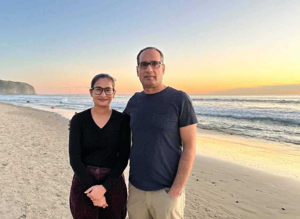 A woman in glasses and a black top stands beside a man in a navy t-shirt on a beach at sunset