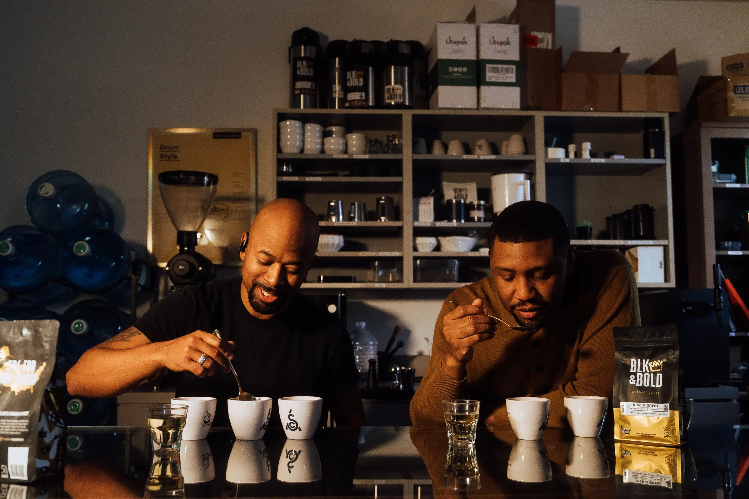 Two men tasting coffee with spoons at a cupping table, with white cups in front of them and a BLK & Bold coffee bag on the right